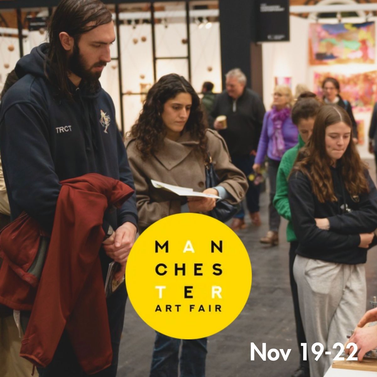 A group of people walking and talking inside a Manchester Art Fair venue, with a large yellow circular logo reading 'MANCHESTER ART FAIR' and the text 'Nov 19-22' overlaid on the image.